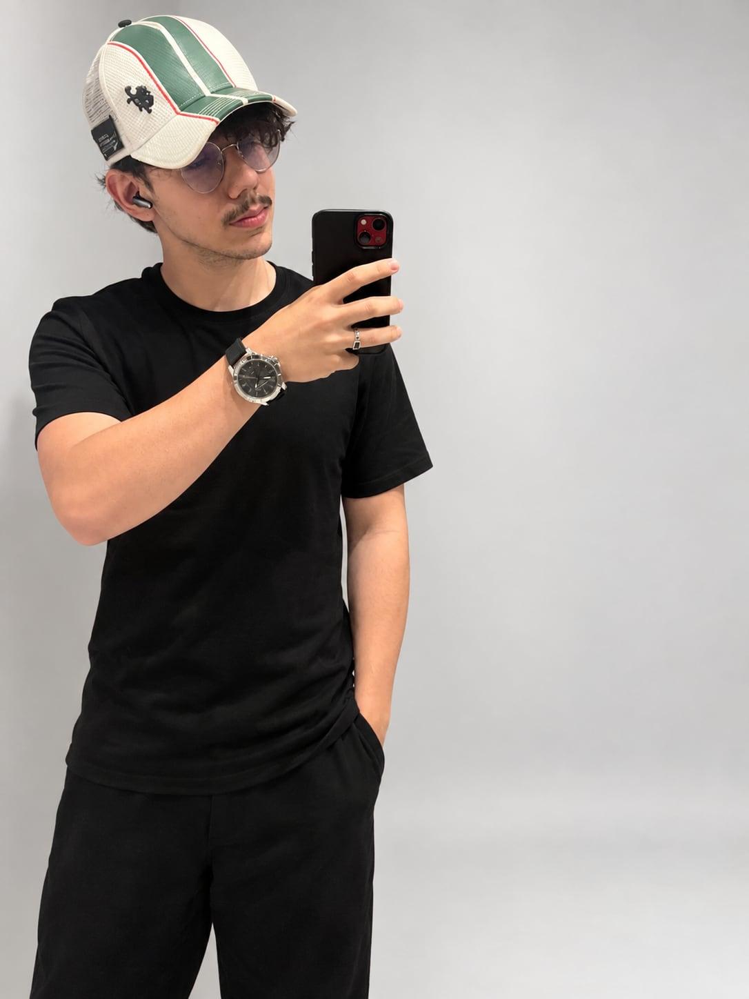 Miguel Eustáquio diante de um espelho, de camiseta preta, boné branco e verde e óculos redondos, fotografando com o celular.